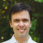 Headshot of a smiling man with short dark hair wearing a white collared shirt, photographed outdoors with a blurred green foliage background.