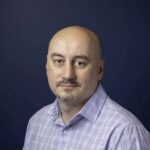 Professional headshot of a man with a mustache wearing a light purple checkered shirt against a dark blue studio background.