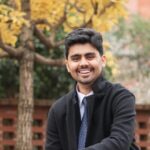 Portrait of a smiling man wearing a black jacket and shirt, standing outdoors with trees and autumn foliage in the background.