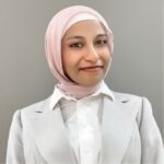 Professional headshot of a woman wearing a light pink hijab and a light gray blazer over a white blouse, smiling against a neutral background.