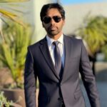 Professional headshot of a man wearing a dark suit, white shirt, and tie, with sunglasses, standing outdoors with greenery and palm trees blurred in the background.