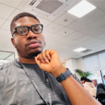 Headshot of a man wearing glasses and a gray shirt, resting his chin on his hand while sitting in an office with ceiling lights and desks in the background.
