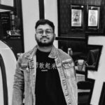 Black-and-white photo of a man wearing glasses, a denim jacket, and a graphic T-shirt, standing indoors with framed posters and equipment visible in the background.