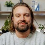 Headshot of a man with shoulder-length hair and a beard, wearing a light gray sweater, seated indoors with plants and shelves in the background.