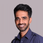 Professional headshot of a smiling man with dark hair and a trimmed beard wearing a dark button-up shirt against a neutral background.