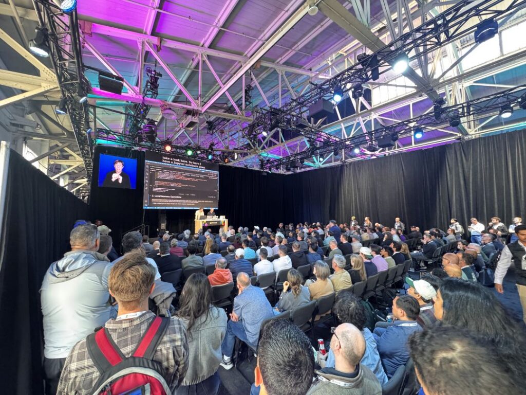 Large audience attending a technical presentation at Microsoft Build 2026. Hundreds of attendees are seated and standing in a packed theater-style session area, watching speakers present on stage beneath large display screens showing live demos and presentation content. Overhead lighting rigs and colorful purple and blue event lighting illuminate the venue, creating an energetic conference atmosphere as developers and technology professionals gather to learn about the latest innovations.