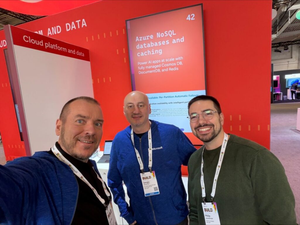 Three Microsoft experts—Marko Hotti, Sergiy Smyrnov, and Phillip Laussermair—pose for a selfie at the Azure NoSQL Databases and Caching booth during Microsoft Build 2026. Behind them is a large digital display promoting Azure NoSQL databases and caching solutions, including Azure Cosmos DB, Azure DocumentDB, and Redis for AI applications. The booth features Microsoft's red event branding and highlights technologies used to power scalable AI and cloud-native applications.
